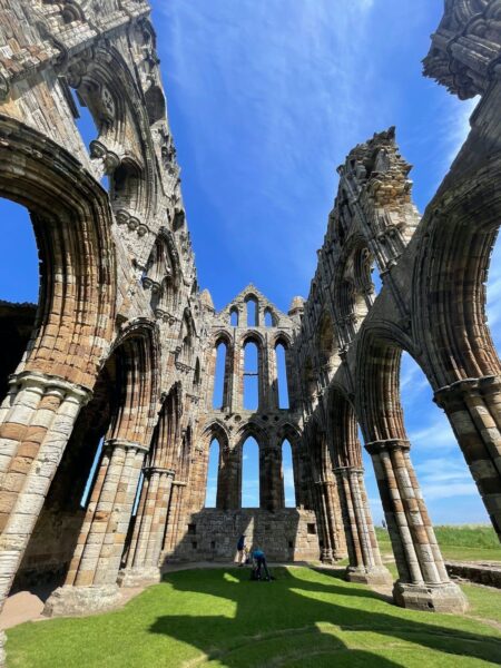 A person sitting on the grass in front of the ruins of an ancient gothic-style abbey with tall arches and clear blue sky in the background.