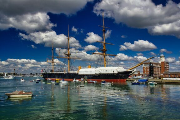 A multi-masted historical tall ship is docked in a harbor, surrounded by smaller boats under a clear sky.