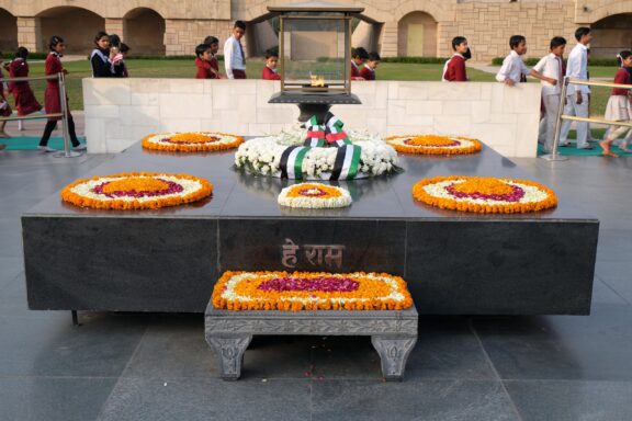 A memorial site with floral wreaths and an eternal flame, surrounded by visitors, located in Raj Ghat, New Delhi.