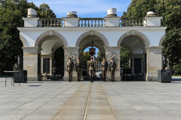 A photo shows a ceremonial guard at Warsaw's Tomb of the Unknown Soldier, surrounded by statues, greenery, and clear skies.