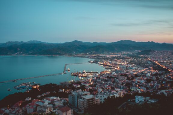 Twilight in Béjaïa, Algeria, with city lights brightening the coastal cityscape, Mediterranean Sea backdrop, and mountain horizon.