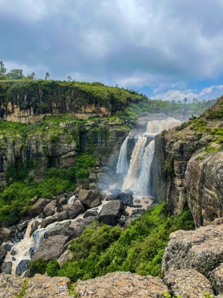 A scenic view of a waterfall at Debre Libanos, Ethiopia, with lush greenery surrounding the cascading water and a cloudy sky above.