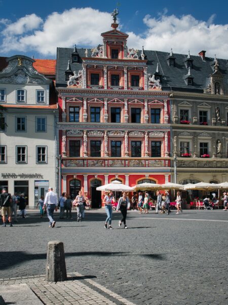 A scenic street view in Erfurt, Germany, featuring pedestrians and a historic red and white building under a cloudy blue sky.