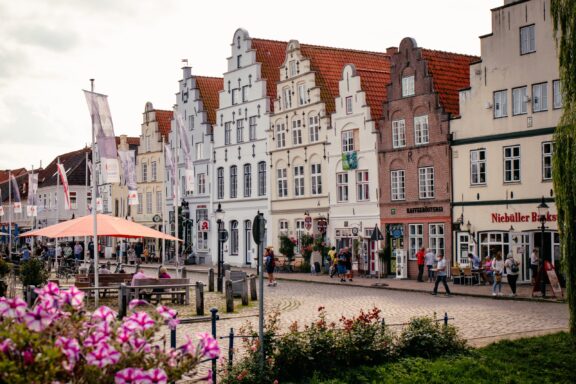 Charming European Town Square with Traditional Facades in Friedrichstadt, Germany A German street view with traditional buildings, pedestrians, a café, and flowers adding a natural touch to the urban scene.