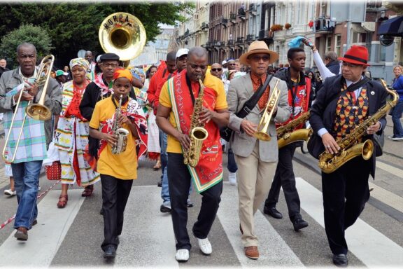 People in colorful attire playing brass and woodwind instruments in a parade, likely for Keti Koti in Suriname.