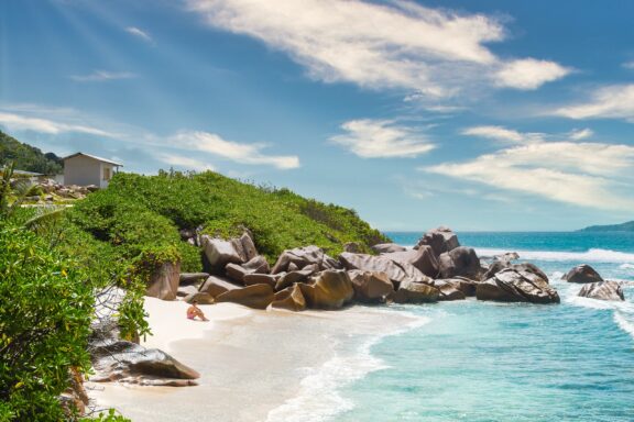 A picturesque view of La Digue in Seychelles featuring a white sandy beach with large granite boulders, lush greenery, and a clear blue sky.