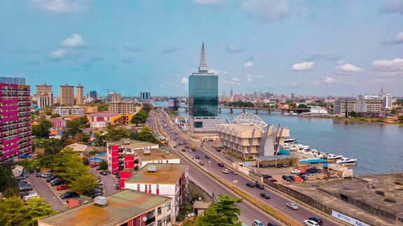 Aerial view of a cityscape with modern buildings, a bridge over a body of water, and vehicles on the road, likely in Lagos, Nigeria.