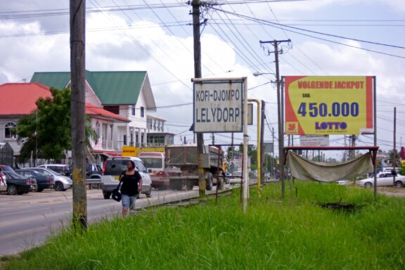 A Lelydorp street scene features a town sign, lottery ad, a cyclist, parked cars, colorful buildings, and a cloudy sky.