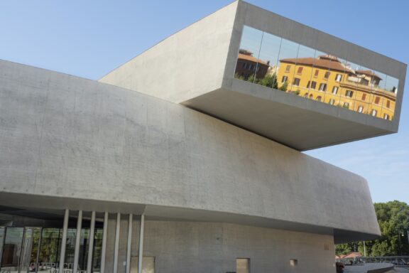 A contemporary building, possibly Rome's MAXXI Art Museum, features concrete slabs, large windows, and reflects its urban setting.