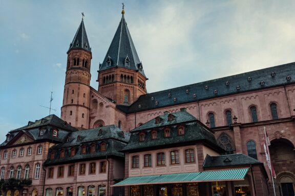 Twilight View of Mainz Cathedral with Adjacent Buildings A photo shows Mainz Cathedral's dual spires and detailed facade against a twilight sky, with nearby buildings slightly visible.