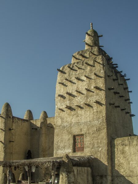 A traditional mud-brick mosque with a distinctive conical tower and wooden beams protruding from the walls, located in Mali, under a clear blue sky.