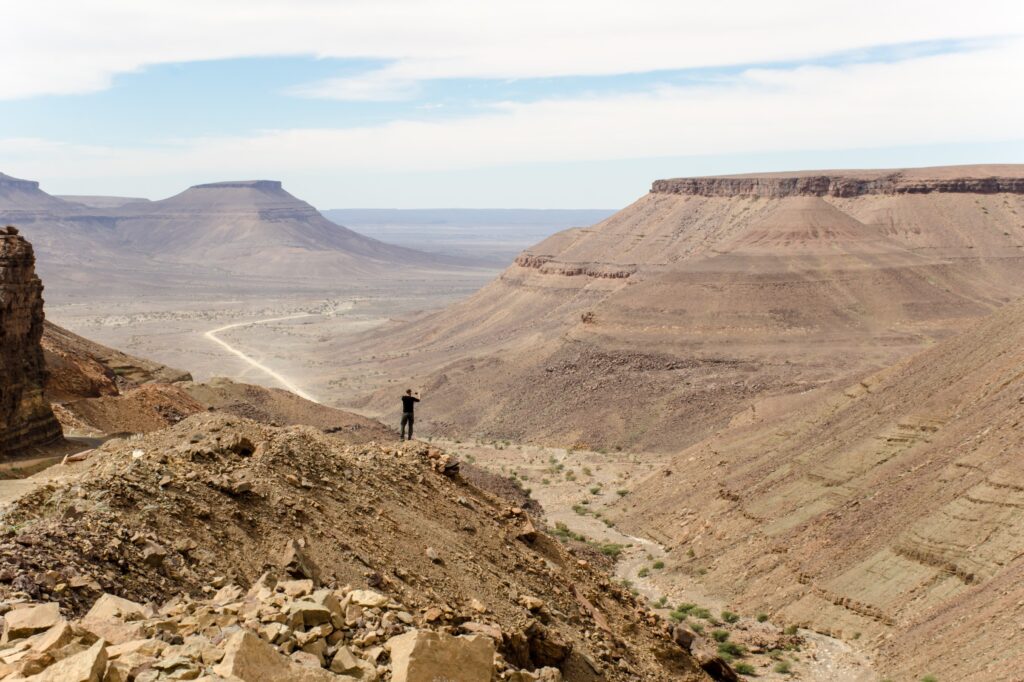 A person standing on a rocky outcrop overlooking a vast, arid landscape with flat-topped mountains and a winding road in Mauritania.