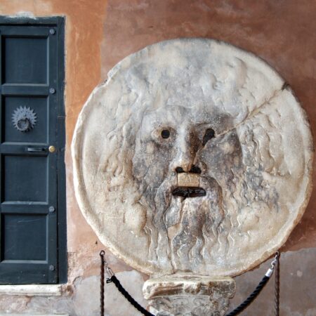 The Mouth of Truth is a round stone sculpture with a face, featuring open eyes and a mouth hole, mounted on a wall.