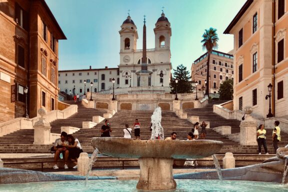 A photo of Piazza di Spagna in Rome with the Spanish Steps, a fountain in the foreground, and people sitting and walking around.