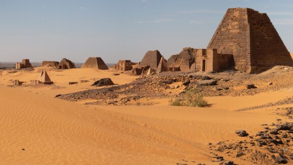 A desert landscape featuring several small pyramids of the Pyramids of Meroë in Sudan, with clear skies above and sand dunes in the foreground.