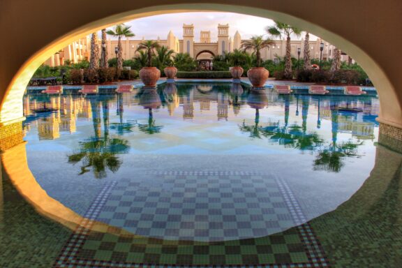 A serene pool view at Riu Touareg Hotel in Cape Verde, framed by an archway with palm tree and architectural reflections.