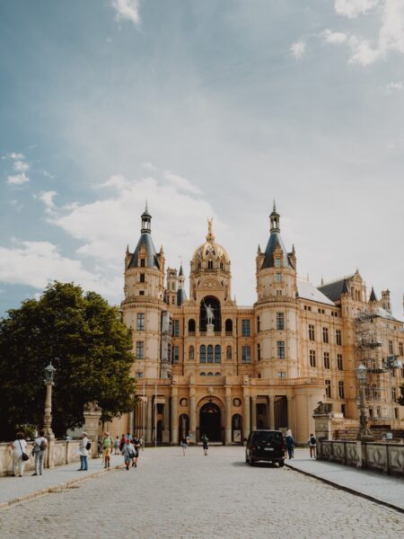 An image of Schwerin Castle in Germany, with people walking in front and a car driving by on a sunny day with a few clouds in the sky.