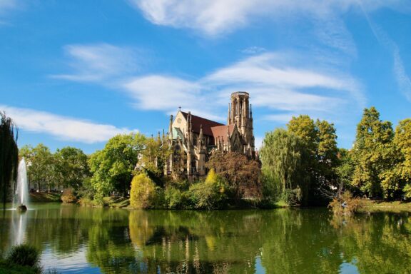 A serene view of Feuersee in Stuttgart, featuring a reflection of a church and surrounding trees on the water's surface under a clear blue sky.