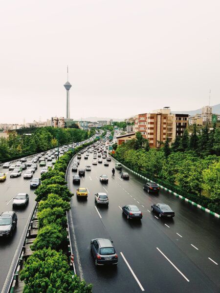 A busy Tehran highway scene with cars, greenery, and the distant Milad Tower under an overcast sky.