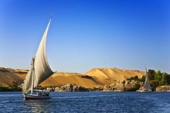 A traditional sailboat, known as a felucca, on the Nile River with sand dunes in the background, under a clear blue sky in Egypt.