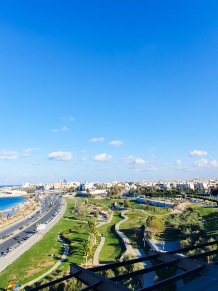 Aerial view of a coastal city park in Tripoli, Libya, with green spaces, pathways, and a road alongside the sea under a clear blue sky.