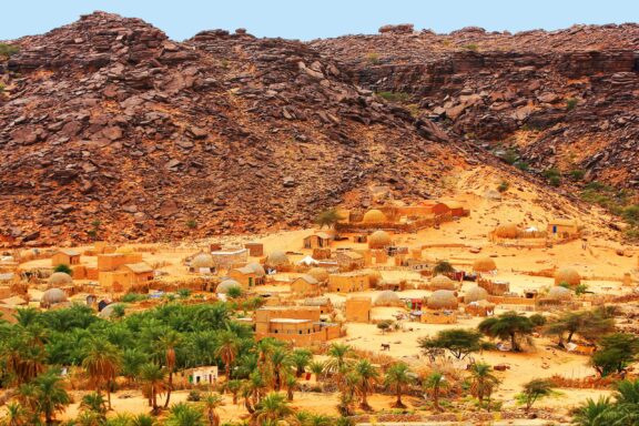 A traditional village with mudbrick buildings lies at a rocky hill's base in the Mhaireth Oasis, Adrar Plateau, Mauritania.