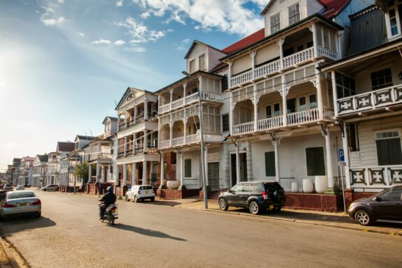 A Paramaribo, Suriname street scene showcases colonial architecture, multi-story buildings, wooden balconies, parked cars, and a motorcyclist.