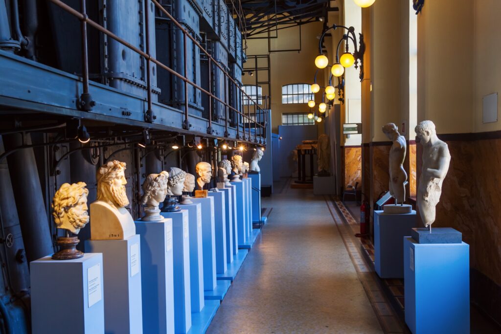 A collection of ancient statues and busts displayed on pedestals inside a room with industrial-style architecture, possibly in a museum in Rome.