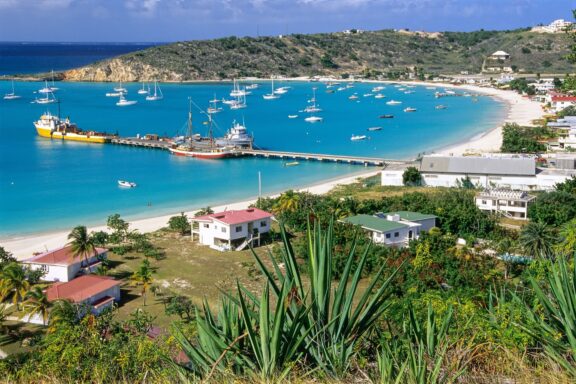 A picturesque coastal scene in Anguilla featuring boats in a clear blue bay, framed by greenery and shoreline buildings.