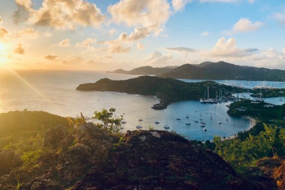 A scenic view of Antigua and Barbuda at sunset, featuring a bay with numerous boats and lush green hills in the background.