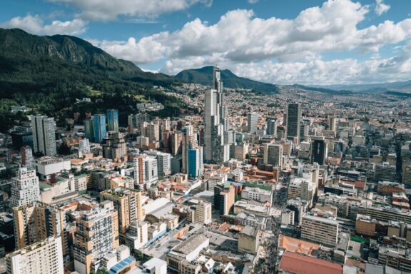 Aerial view of Bogotá, Colombia displays dense urban architecture, skyscrapers, and mountains under a partly cloudy sky.