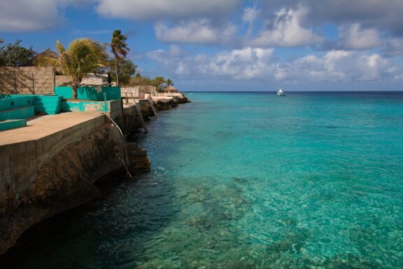 A coastal view in Bonaire featuring turquoise waters, a stone barrier, buildings, greenery, and a partly cloudy sky.