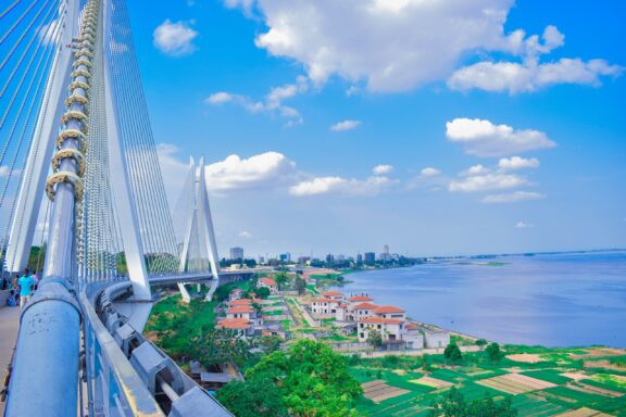 View from a suspension bridge, featuring cables, a river, distant cityscape under a blue sky, possibly in Brazzaville or Kinshasa.