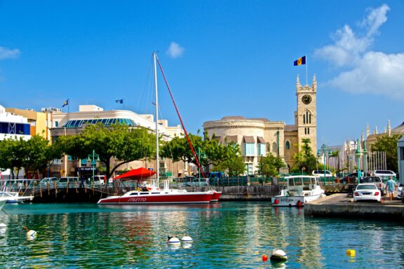 A scenic view of Bridgetown, Barbados, featuring a calm marina with moored boats, clear blue skies, and colorful buildings along the waterfront.