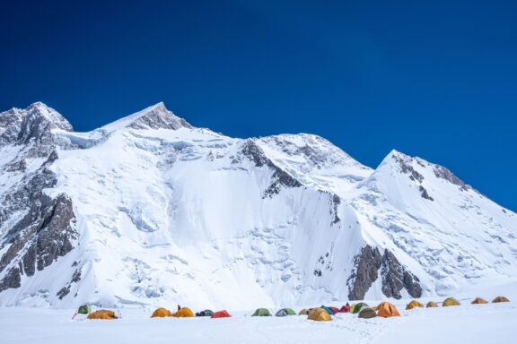 A base camp with colorful tents set against the backdrop of the snow-covered Gasherbrum II mountain.