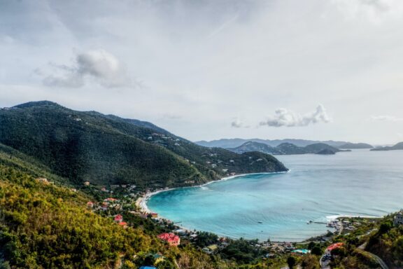 Cane Garden Bay on Tortola Island offers a picturesque view of green hills, a turquoise bay, beachfront, boats, and a partly cloudy sky.