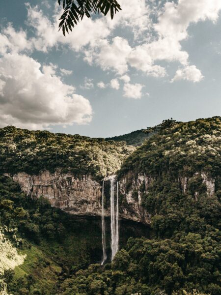 A picturesque view of Brazil's Caracol Waterfall, a tall cascade amidst a green cliff and forest under a semi-cloudy sky.