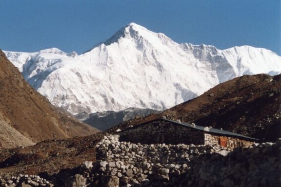 A view of Cho Oyu mountain from Gokyo with a snow-covered peak towering over rocky terrain and a suspension bridge in the foreground.