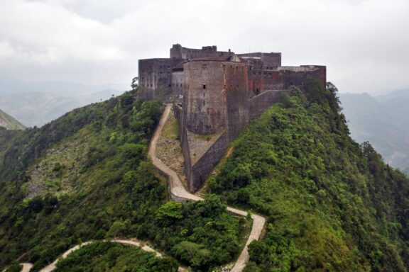 Aerial view of the historic Citadel La Ferriere, a large mountaintop fortress in Haiti, surrounded by greenery with a cloudy sky overhead.