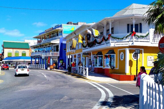 A vibrant street scene in George Town, Cayman Islands, featuring colorful buildings, a clear blue sky, and a few pedestrians and vehicles.