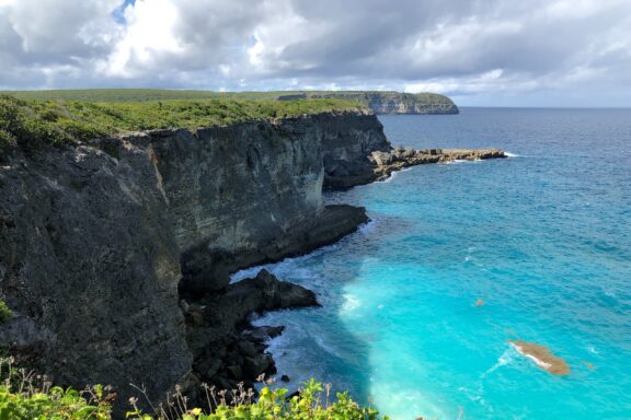 A coastal cliffside view of Grande-Terre, Guadeloupe, with turquoise waters below and a partly cloudy sky above.