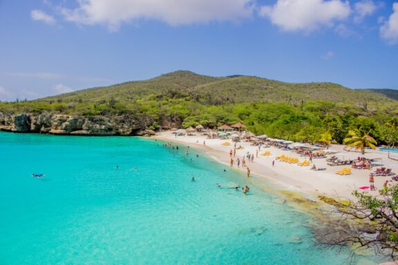 Grote Knip beach in Curacao boasts turquoise waters, a sandy beach with sunbathers, colorful umbrellas, greenery and cliffs.