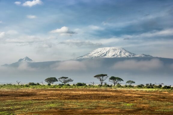 Mount Kilimanjaro's snowy peak towers above the clouds, viewed from Kenya's Amboseli Park, amidst a savannah landscape.