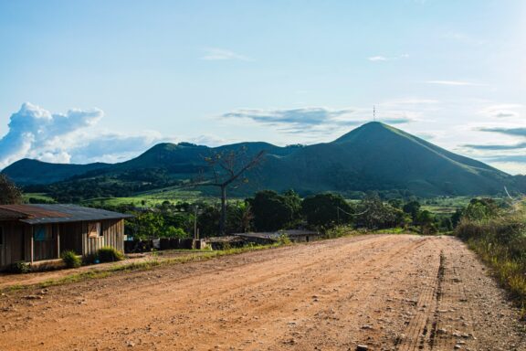 A dirt road in Lope-Ogooue Ivindo, Gabon, leads to green hills under a cloudy blue sky, with small buildings on the left.