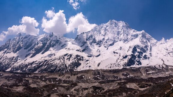 A panoramic view of the snow-capped Manaslu mountain range under a blue sky with scattered clouds.