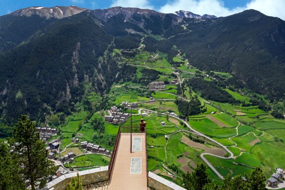 Mirador Roc del Quer in Andorra offers a scenic view of a green valley, village, and mountains, with a wooden platform for visitors.