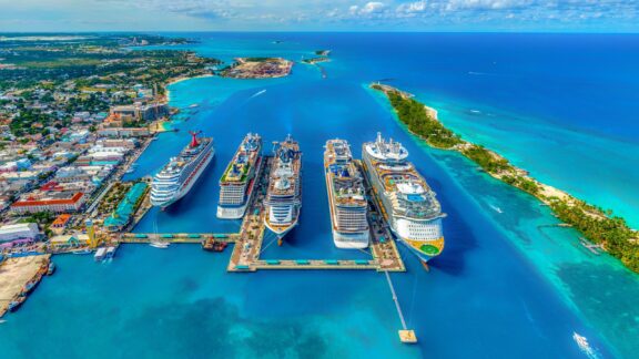 Aerial view of Nassau, Bahamas, showcasing multiple cruise ships docked at a pier with clear blue waters and urban landscape in the background.