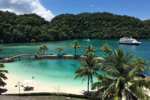 A tropical Palau scene with a turquoise lagoon, lush greenery, a bridge to an islet, palm trees, and distant anchored boats.