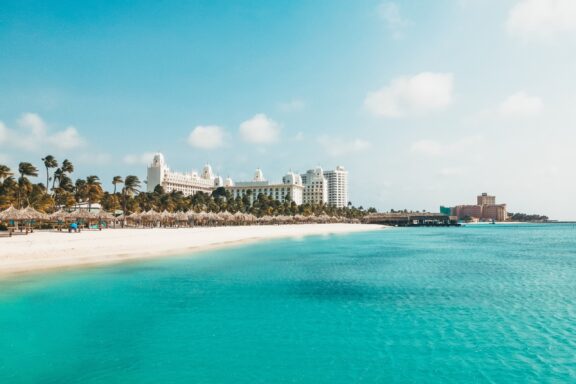 A scenic view of Palm Beach in Aruba, featuring clear turquoise waters, a white sandy beach, and a row of resort buildings under a bright blue sky.