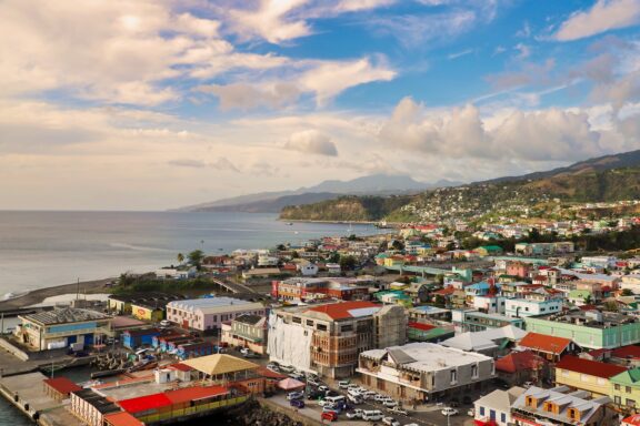 Aerial view of Roseau, Dominica, featuring colorful buildings, a coastline with calm sea, and hills in the background under a partly cloudy sky.